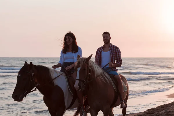 Una pareja amorosa en ropa de verano montando un caballo en una playa de arena al atardecer. Mar y puesta de sol en el fondo. Enfoque selectivo —  Fotos de Stock