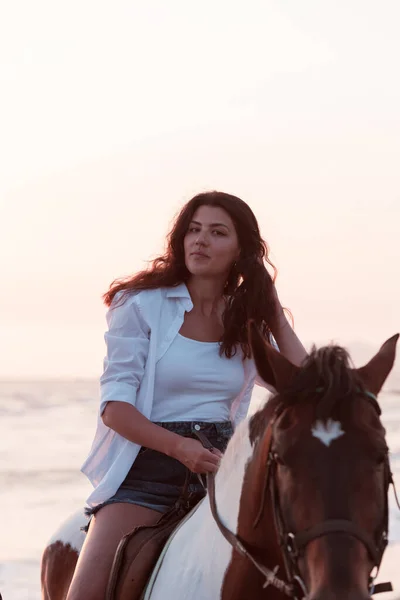 Woman in summer clothes enjoys riding a horse on a beautiful sandy beach at sunset. Selective focus — Zdjęcie stockowe