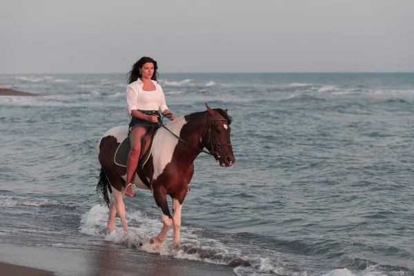 Mujer en ropa de verano disfruta montar a caballo en una hermosa playa de arena al atardecer. Enfoque selectivo —  Fotos de Stock