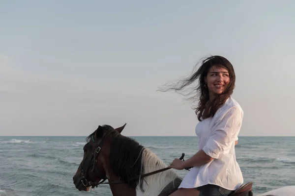 Woman in summer clothes enjoys riding a horse on a beautiful sandy beach at sunset. Selective focus — Fotografia de Stock