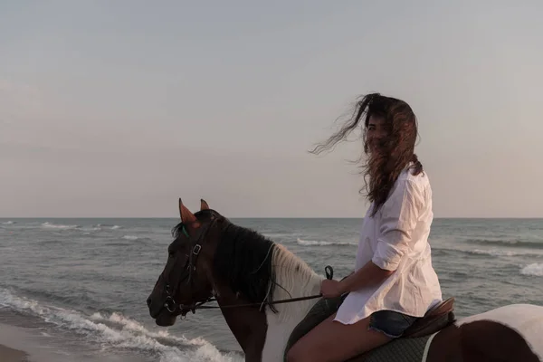 Woman in summer clothes enjoys riding a horse on a beautiful sandy beach at sunset. Selective focus — Stock Fotó