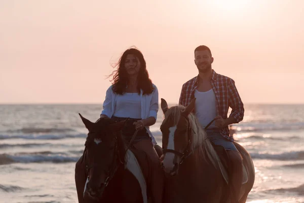 Gün batımında kumlu bir sahilde at süren, yaz kıyafetleri içinde sevgi dolu genç bir çift. Arka planda deniz ve gün batımı. Seçici odak — Stok fotoğraf