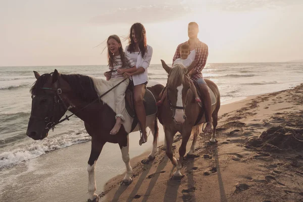 The family spends time with their children while riding horses together on a beautiful sandy beach on sunet. — Zdjęcie stockowe