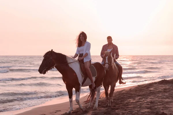 Любляча молода пара в літньому одязі катається на коні на піщаному пляжі на заході сонця. Море і захід сонця на задньому плані. Вибірковий фокус — стокове фото