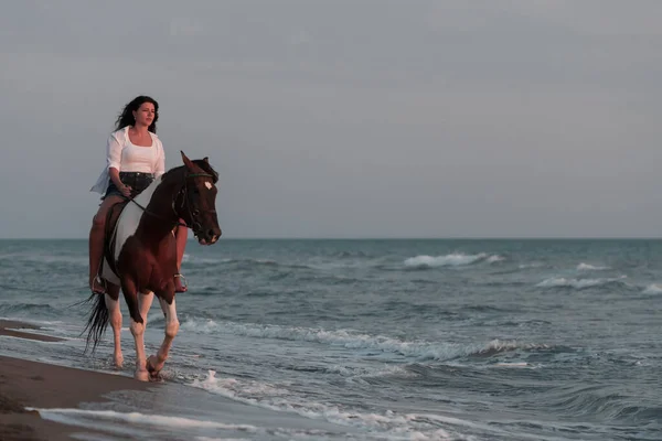 Mujer en ropa de verano disfruta montar a caballo en una hermosa playa de arena al atardecer. Enfoque selectivo —  Fotos de Stock