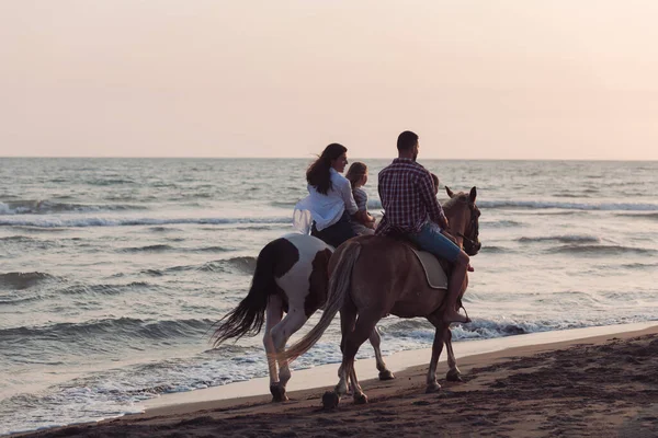 The family spends time with their children while riding horses together on a beautiful sandy beach on sunet. — Fotografia de Stock