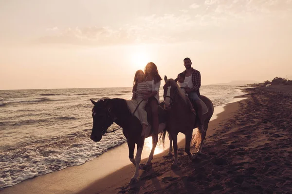 The family spends time with their children while riding horses together on a beautiful sandy beach on sunet. — Zdjęcie stockowe