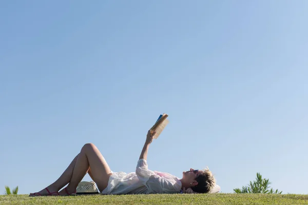 Kvinna ljuger och läser sin favoritbok på en äng täckt med färskt grönt gräs en solig sommar eller vår dag. — Stockfoto