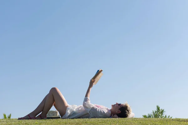 Mujer acostada y leyendo su libro favorito en un prado cubierto de hierba verde fresca en un soleado día de verano o primavera. — Foto de Stock