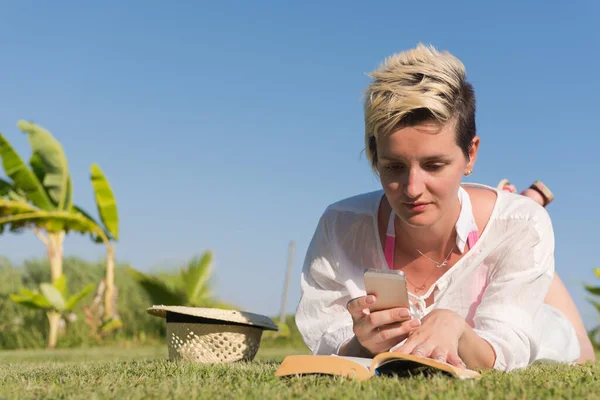 Kvinna ljuger och läser sin favoritbok på en äng täckt med färskt grönt gräs en solig sommar eller vår dag. — Stockfoto