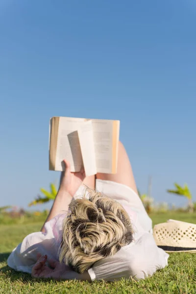 Kvinna ljuger och läser sin favoritbok på en äng täckt med färskt grönt gräs en solig sommar eller vår dag. — Stockfoto