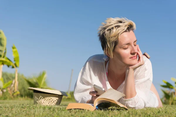 女性が横たわって、晴れた夏や春の日に新鮮な緑の草で覆われた牧草地で彼女の好きな本を読んで. — ストック写真