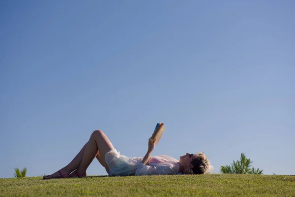 Kvinna ljuger och läser sin favoritbok på en äng täckt med färskt grönt gräs en solig sommar eller vår dag. — Stockfoto