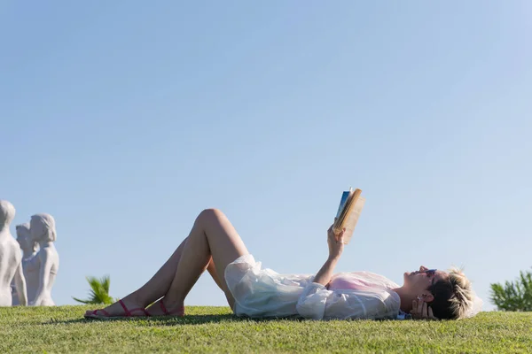 女性が横たわって、晴れた夏や春の日に新鮮な緑の草で覆われた牧草地で彼女の好きな本を読んで. — ストック写真