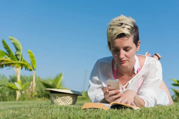 Kvinna ljuger och läser sin favoritbok på en äng täckt med färskt grönt gräs en solig sommar eller vår dag. — Stockfoto