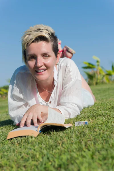 Frau liegt und liest ihr Lieblingsbuch auf einer mit frischem grünen Gras bedeckten Wiese an einem sonnigen Sommer- oder Frühlingstag. — Stockfoto