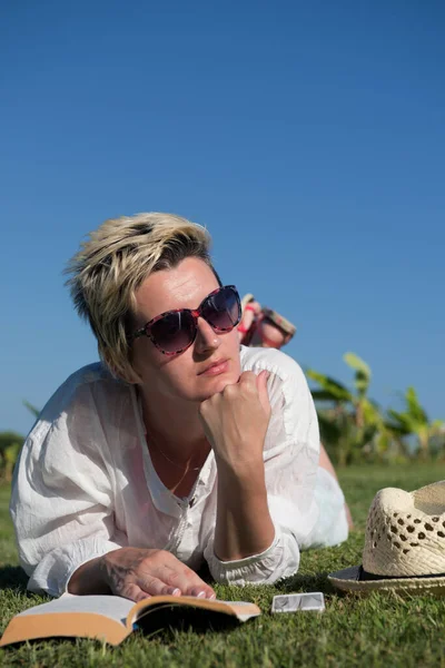 Frau liegt und liest ihr Lieblingsbuch auf einer mit frischem grünen Gras bedeckten Wiese an einem sonnigen Sommer- oder Frühlingstag. — Stockfoto