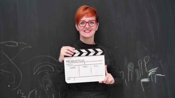 영화 클리퍼 보드를 들고 웃으며 분필 보드 배경에 카메라를 보고 있는 빨간 머리 여자 배우. — 비디오