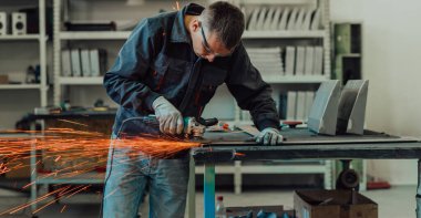 Heavy Industry Engineering Factory Interior with Industrial Worker Using Angle Grinder and Cutting a Metal Tube. Güvenlik Üniforması ve Sert Şapka Üretim Metal Yapıları Müteahhiti. Yüksek kalite