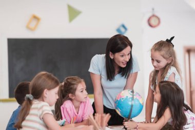 Female teacher with kids in geography class looking at a globe. Side view of a group of diverse happy school kids with a globe in the classroom at school. High quality photo