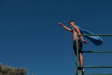 young teen boy near the river having fun with friends at a summer party wearing towel as a superhero scarf ready for a jump looking brave in distance. High quality photo