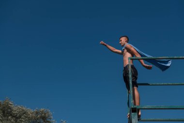 young teen boy near the river having fun with friends at a summer party wearing towel as a superhero scarf ready for a jump looking brave in distance. High quality photo