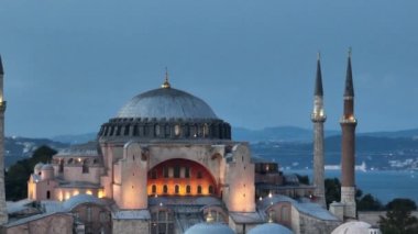 Establishing orbiting aerial drone shot of a Hagia Sophia Holy Grand Mosque with Bosphorus bridge and city skyline with a flag on the background in Fatih, Istanbul, Turkey at sunset. Cinematic footage