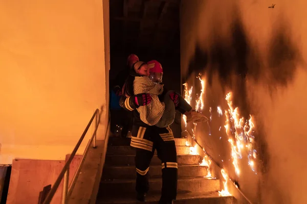Brave Fireman Descends Stairs of a Burning Building and Holds Saved Girl in His Arms. Open fire and one Firefighter in the Background. High quality photo