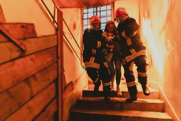 Brave Fireman Descends Stairs of a Burning Building and Holds Saved Girl in His Arms. Open fire and one Firefighter in the Background. High quality photo