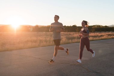 Sağlıklı genç bir çift sabahın erken saatlerinde arka planda güzel bir gündoğumuyla şehir sokaklarında koşuyor. Yüksek kalite fotoğraf