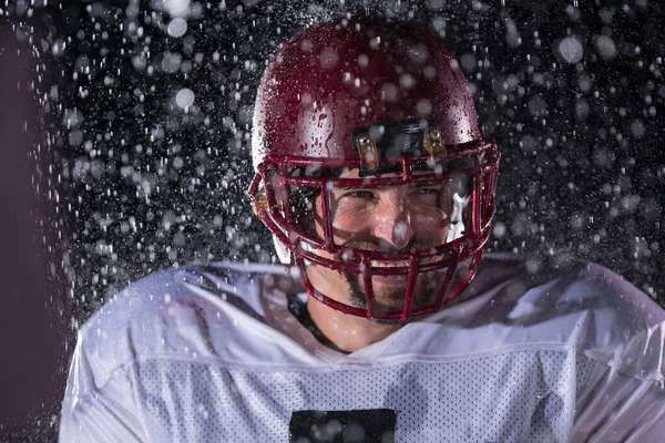 American Football Athlete Warrior Standing on a Field Holds his Helmet and Ready to Play. Player Preparing to Run, Attack and Score Touchdown. Rainy Night with Dramatic lens flare and rain drops. High