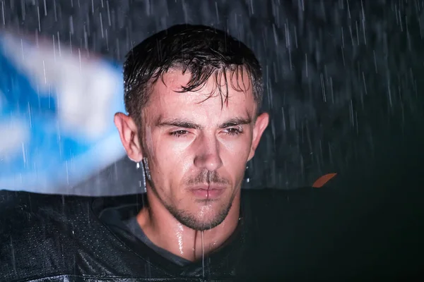 American Football Athlete Warrior Standing on a Field Holds his Helmet and Ready to Play. Player Preparing to Run, Attack and Score Touchdown. Rainy Night with Dramatic lens flare and rain drops. High quality photo