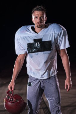 Sahada Ayakta Duran Amerikan Futbolu Sporcusu Miğferini Tutuyor ve Oynamaya Hazır. Oyuncu koşmaya, saldırmaya ve sayı yapmaya hazırlanıyor. Dramatik mercekler ve yağmur damlalarıyla dolu yağmurlu bir gece. Yüksek