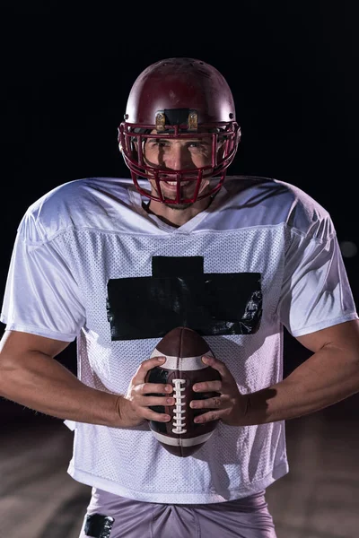 American Football Athlete Warrior Standing on a Field Holds his Helmet and Ready to Play. Player Preparing to Run, Attack and Score Touchdown. Rainy Night with Dramatic lens flare and rain drops. High