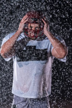 Sahada Ayakta Duran Amerikan Futbolu Sporcusu Miğferini Tutuyor ve Oynamaya Hazır. Oyuncu koşmaya, saldırmaya ve sayı yapmaya hazırlanıyor. Dramatik mercekler ve yağmur damlalarıyla dolu yağmurlu bir gece. Yüksek