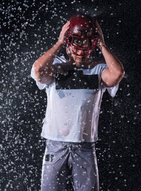 Sahada Ayakta Duran Amerikan Futbolu Sporcusu Miğferini Tutuyor ve Oynamaya Hazır. Oyuncu koşmaya, saldırmaya ve sayı yapmaya hazırlanıyor. Dramatik mercekler ve yağmur damlalarıyla dolu yağmurlu bir gece. Yüksek