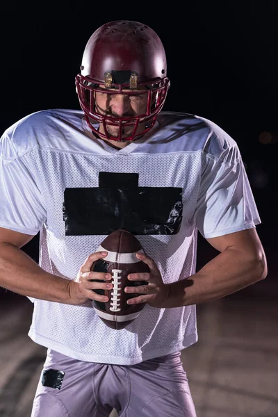 American Football Athlete Warrior Standing on a Field Holds his Helmet and Ready to Play. Player Preparing to Run, Attack and Score Touchdown. Rainy Night with Dramatic lens flare and rain drops. High