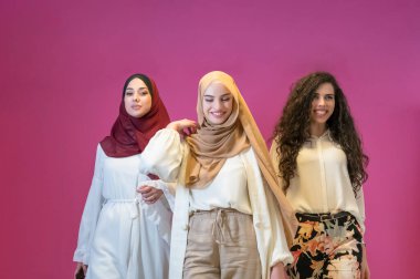 Modern giysiler içinde üç Müslüman kadın pembe arka planda poz veriyor. 
