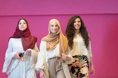 Modern giysiler içinde üç Müslüman kadın pembe arka planda poz veriyor. 