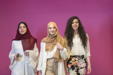 Modern giysiler içinde üç Müslüman kadın pembe arka planda poz veriyor. 