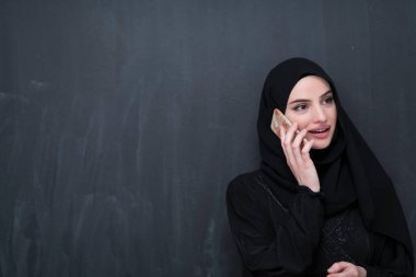 Genç, modern Müslüman iş kadınları siyah bir karatahtanın önünde tesettürlü kıyafetler giyerek akıllı telefon kullanıyorlar..