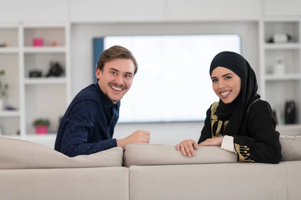 若いイスラム教徒のカップルの女性を身に着けていますイスラムヒジャーブ服を身に着けていますソファに座ってテレビを見ます一緒にラマダーン月の間に現代的な家で — ストック写真
