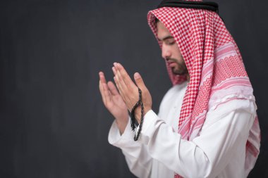 Geleneksel kıyafetlerle Tanrı 'ya dua eden genç bir Arap adam ellerini siyah bir arkaplan önünde dua ederek tutar. Yüksek kalite fotoğraf