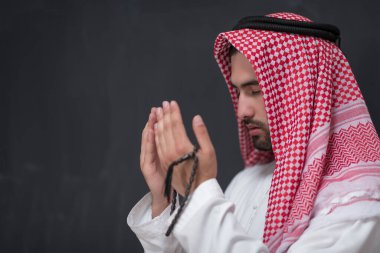 Geleneksel kıyafetlerle Tanrı 'ya dua eden genç bir Arap adam ellerini siyah bir arkaplan önünde dua ederek tutar. Yüksek kalite fotoğraf