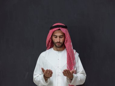 Geleneksel kıyafetlerle Tanrı 'ya dua eden genç bir Arap adam ellerini siyah bir arkaplan önünde dua ederek tutar. Yüksek kalite fotoğraf