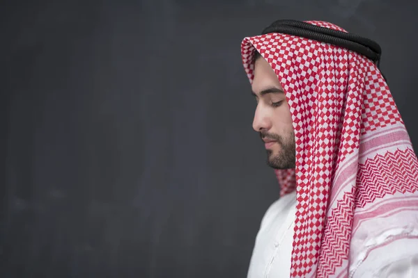 A young Arabian man in traditional clothes making a traditional prayer to God keeps his hands in praying gesture in front of a black background. High quality photo