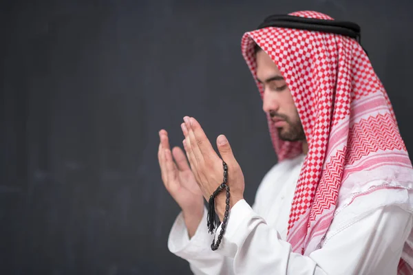 A young Arabian man in traditional clothes making a traditional prayer to God keeps his hands in praying gesture in front of a black background. High quality photo