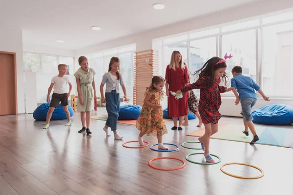 教室内の床の上に女性教師がいる小さな保育園の子供たちは 運動をしています フラフープサークルを飛び越えて床の上を追跡します 高品質の写真 — ストック写真
