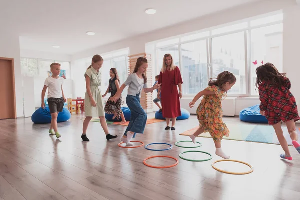 Small nursery school children with a female teacher on the floor indoors in the classroom, doing exercise. Jumping over hula hoop circles track on the floor. High quality photo