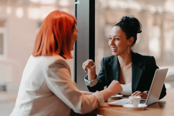 Two young businesswomen sitting at a table in a cafe. The woman shows colleague information on the laptop screen. Girl using smartphone, blogging. Teamwork, business meetings. High quality photo